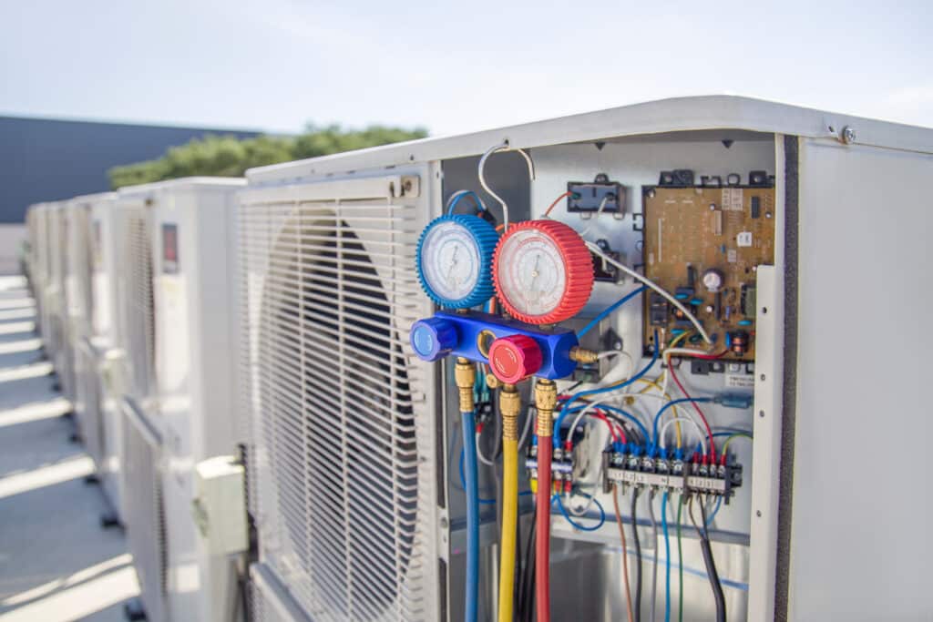 An open outdoor HVAC unit with a manifold gauge set attached, showing exposed wiring and internal components, highlights the importance of professional HVAC services in Palmyra PA, on a rooftop with similar units in the background.